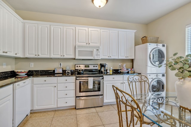 kitchen featuring white cabinetry, stacked washer / drying machine, light tile patterned floors, dark stone counters, and white appliances