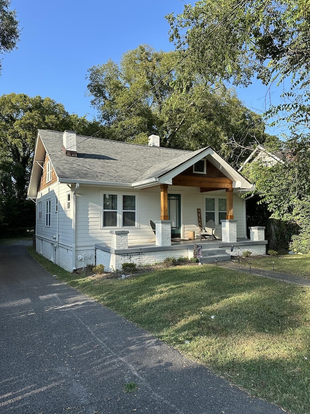 view of front of property featuring covered porch, a chimney, a front yard, and a shingled roof