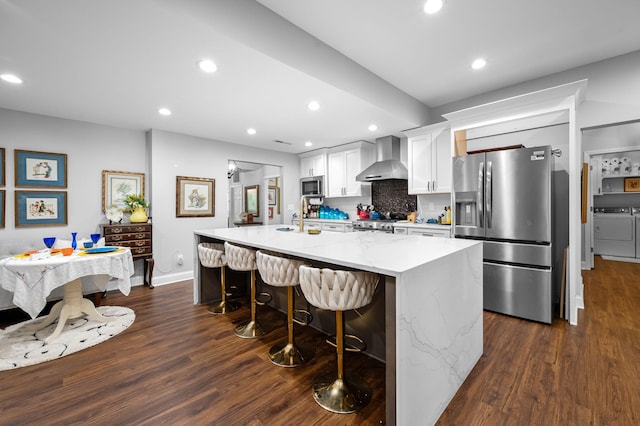 kitchen featuring washer and clothes dryer, dark wood-type flooring, a kitchen island with sink, stainless steel appliances, and wall chimney range hood