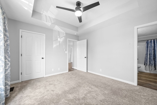 unfurnished bedroom with a tray ceiling, ceiling fan, carpet floors, and ensuite bathroom