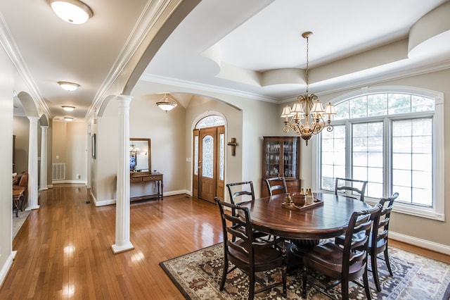 dining room with a raised ceiling, an inviting chandelier, wood-type flooring, ornate columns, and crown molding