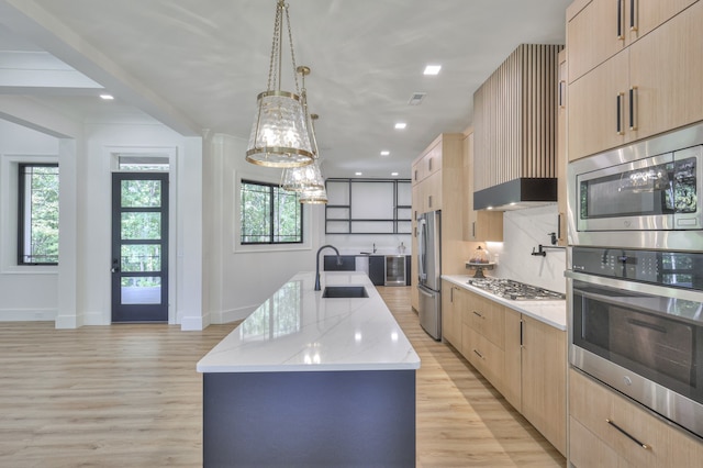 kitchen with pendant lighting, light hardwood / wood-style flooring, light brown cabinetry, appliances with stainless steel finishes, and a kitchen island with sink