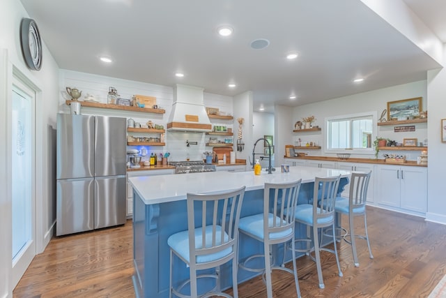 kitchen with white cabinets, hardwood / wood-style floors, stainless steel appliances, premium range hood, and a center island with sink