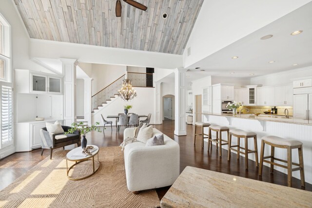 living room featuring dark hardwood / wood-style floors, ceiling fan with notable chandelier, decorative columns, wood ceiling, and high vaulted ceiling