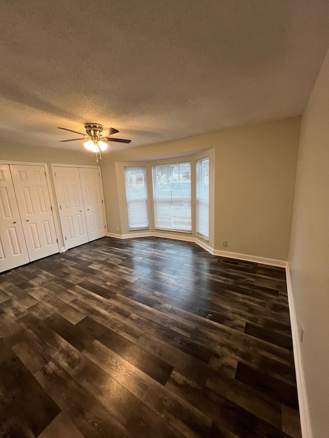 unfurnished bedroom with a textured ceiling, a ceiling fan, dark wood finished floors, baseboards, and two closets