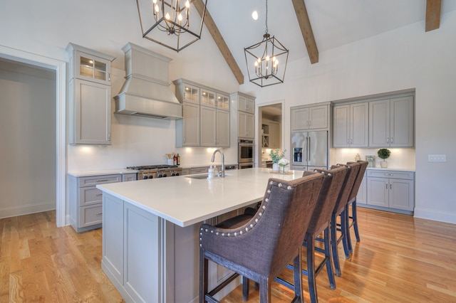 kitchen with custom range hood, appliances with stainless steel finishes, a kitchen island with sink, sink, and a chandelier