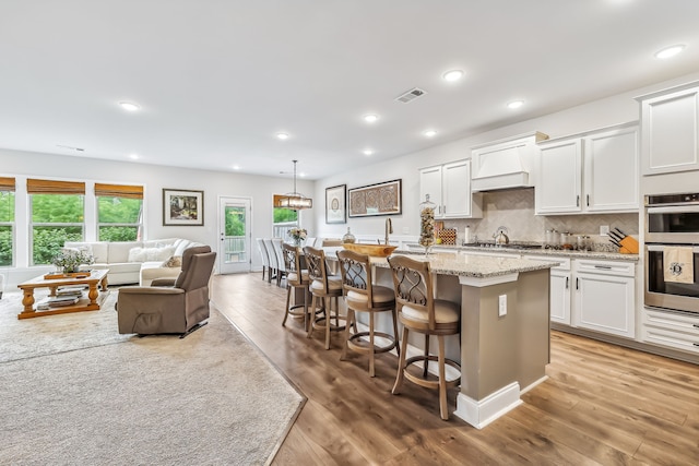 kitchen with white cabinets, pendant lighting, a kitchen island with sink, light wood-type flooring, and decorative backsplash