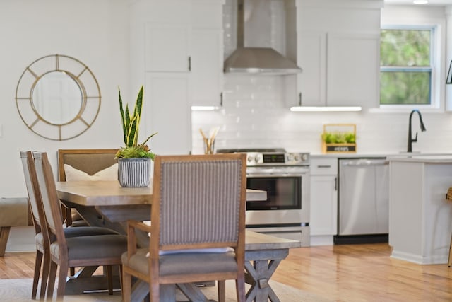 kitchen with wall chimney range hood, appliances with stainless steel finishes, sink, light hardwood / wood-style floors, and white cabinets