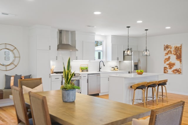 kitchen featuring stainless steel appliances, wall chimney range hood, white cabinets, and hanging light fixtures