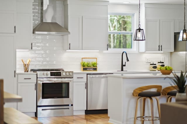 kitchen with stainless steel appliances, white cabinetry, decorative light fixtures, wall chimney exhaust hood, and light hardwood / wood-style flooring