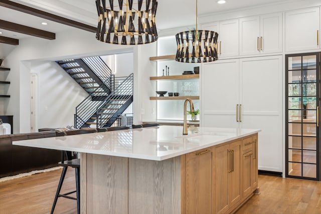 interior space with a kitchen island with sink, light wood-type flooring, white cabinetry, sink, and beam ceiling