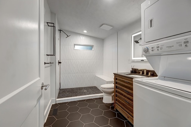 bathroom featuring vanity, a tile shower, stacked washing maching and dryer, tile patterned flooring, and toilet