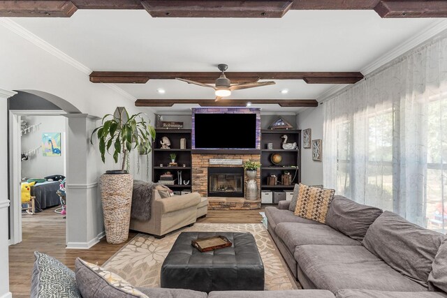 living room with light hardwood / wood-style flooring, a stone fireplace, beam ceiling, crown molding, and ceiling fan