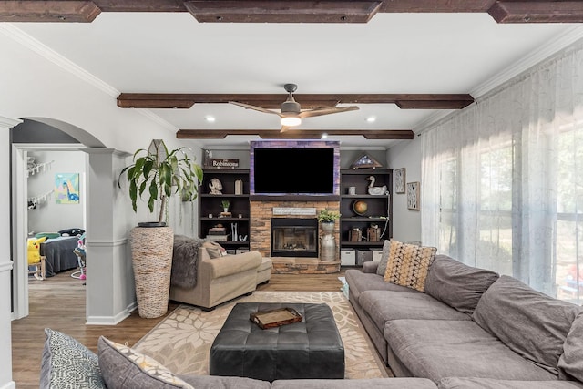 living room featuring a ceiling fan, arched walkways, ornamental molding, a stone fireplace, and light wood-style floors
