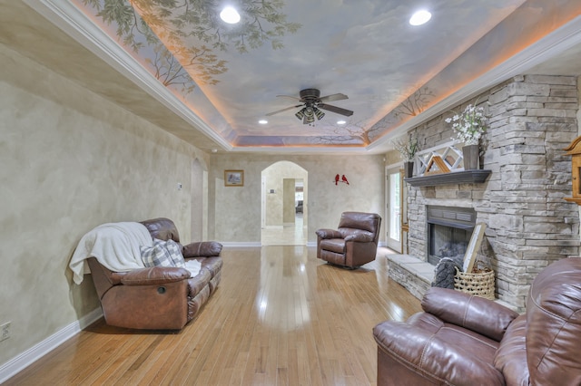 living room featuring light hardwood / wood-style flooring, a stone fireplace, a tray ceiling, ornamental molding, and ceiling fan