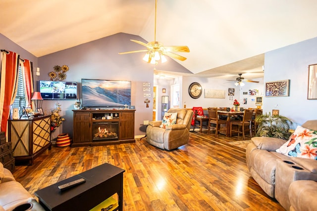 living area with ceiling fan, a warm lit fireplace, wood finished floors, and vaulted ceiling