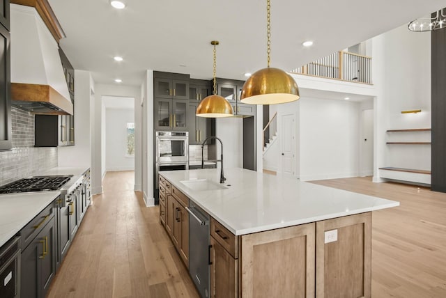 kitchen featuring glass insert cabinets, custom exhaust hood, stainless steel appliances, light wood-type flooring, and a sink