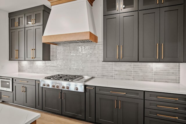 kitchen featuring light wood-style flooring, custom range hood, glass insert cabinets, stainless steel gas cooktop, and backsplash