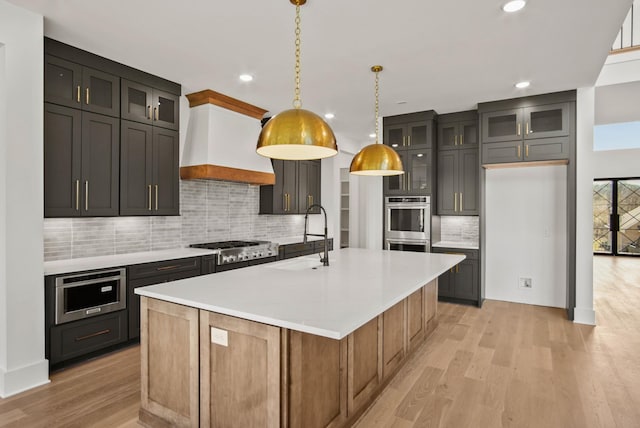 kitchen with light wood finished floors, light countertops, a sink, an island with sink, and premium range hood