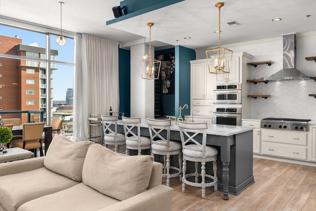 kitchen with hanging light fixtures, light wood-type flooring, wall chimney exhaust hood, and tasteful backsplash