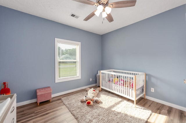 bedroom featuring visible vents, wood finished floors, a nursery area, baseboards, and ceiling fan