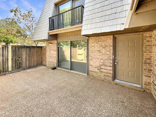 property entrance featuring brick siding, fence, mansard roof, a balcony, and a patio