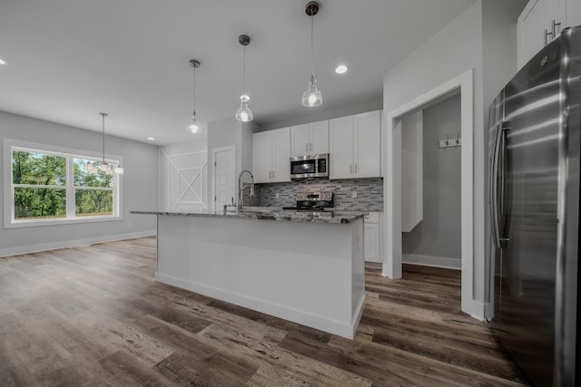 kitchen with decorative light fixtures, appliances with stainless steel finishes, a kitchen island with sink, light stone counters, and white cabinets