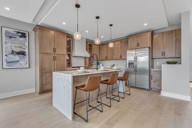 kitchen featuring light wood-type flooring, stainless steel fridge, a kitchen bar, an island with sink, and custom range hood