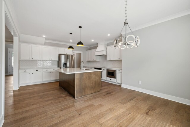 kitchen with custom range hood, appliances with stainless steel finishes, a kitchen island with sink, hanging light fixtures, and hardwood / wood-style flooring