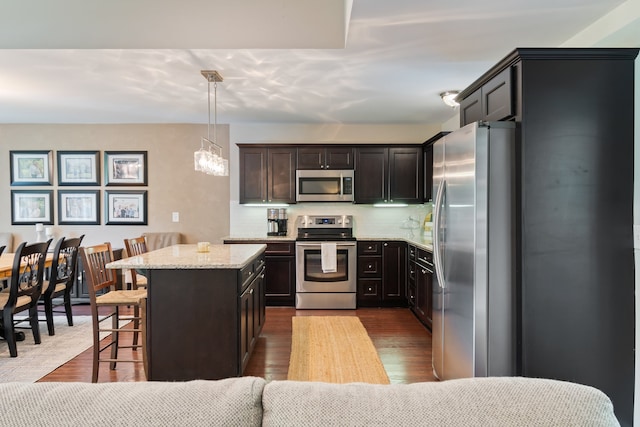 kitchen with hardwood / wood-style floors, appliances with stainless steel finishes, hanging light fixtures, light stone counters, and dark brown cabinetry