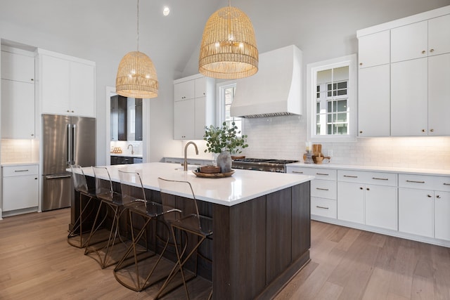 kitchen with custom range hood, appliances with stainless steel finishes, light hardwood / wood-style floors, a kitchen island with sink, and white cabinets