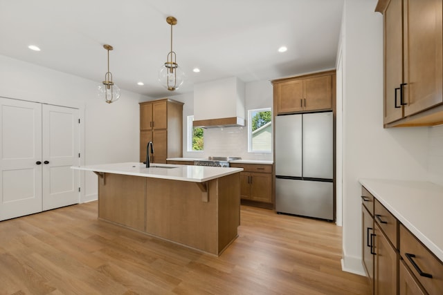 kitchen with hanging light fixtures, light wood-type flooring, stainless steel appliances, premium range hood, and an island with sink