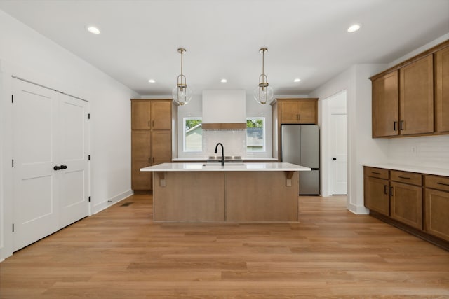 kitchen with light wood-type flooring, a kitchen island with sink, stainless steel fridge, decorative light fixtures, and sink