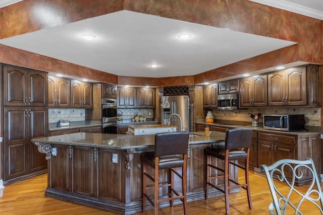 kitchen featuring light hardwood / wood-style flooring, stainless steel appliances, dark stone counters, a breakfast bar area, and a kitchen island with sink