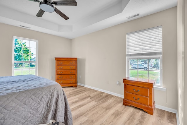 bedroom featuring ceiling fan, a tray ceiling, multiple windows, and light hardwood / wood-style floors