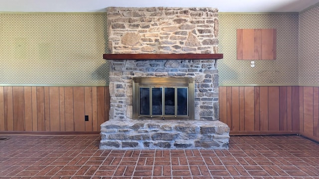 unfurnished living room featuring brick floor, a wainscoted wall, wood walls, a stone fireplace, and wallpapered walls