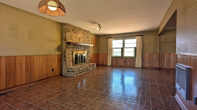 unfurnished living room featuring a wainscoted wall, heating unit, brick patterned floor, a stone fireplace, and wallpapered walls