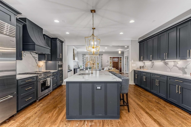 kitchen with pendant lighting, sink, high quality appliances, custom range hood, and a center island with sink