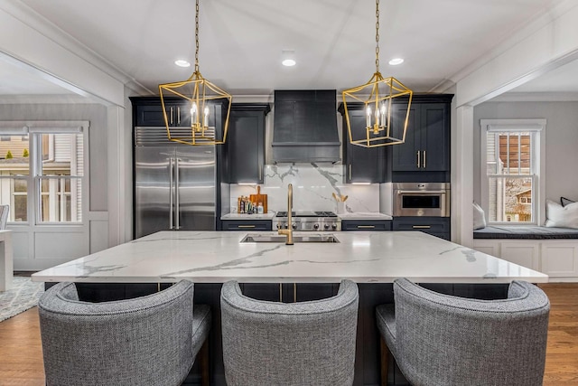 kitchen with custom exhaust hood, an island with sink, pendant lighting, and stainless steel appliances