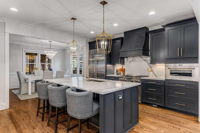 kitchen with high end appliances, pendant lighting, premium range hood, and a center island with sink