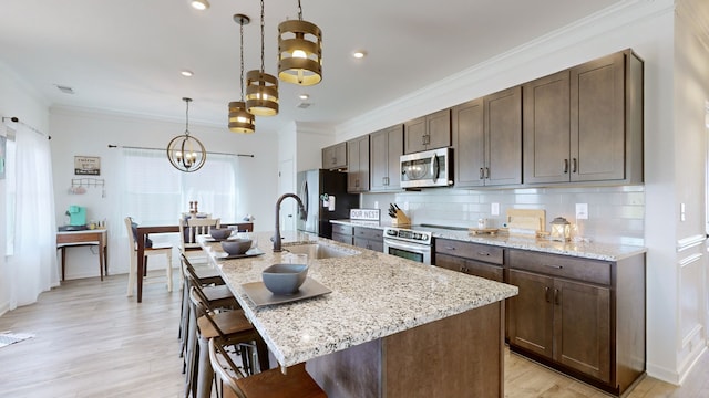 kitchen with pendant lighting, stainless steel appliances, light wood-type flooring, crown molding, and a kitchen island with sink