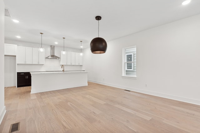 kitchen featuring white cabinets, hanging light fixtures, wall chimney exhaust hood, light wood-type flooring, and an island with sink