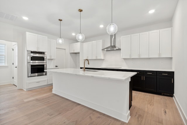 kitchen with sink, wall chimney exhaust hood, stainless steel double oven, an island with sink, and white cabinetry