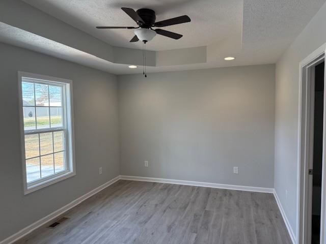 unfurnished room featuring a raised ceiling, ceiling fan, light hardwood / wood-style flooring, and a textured ceiling