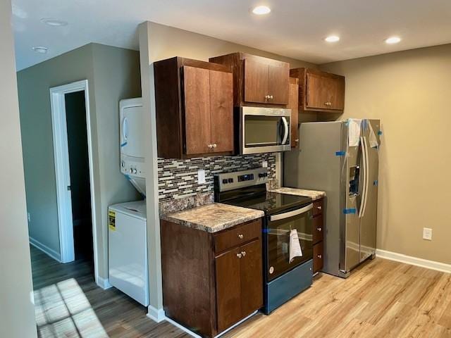 kitchen featuring light wood-style flooring, appliances with stainless steel finishes, baseboards, backsplash, and stacked washer / drying machine