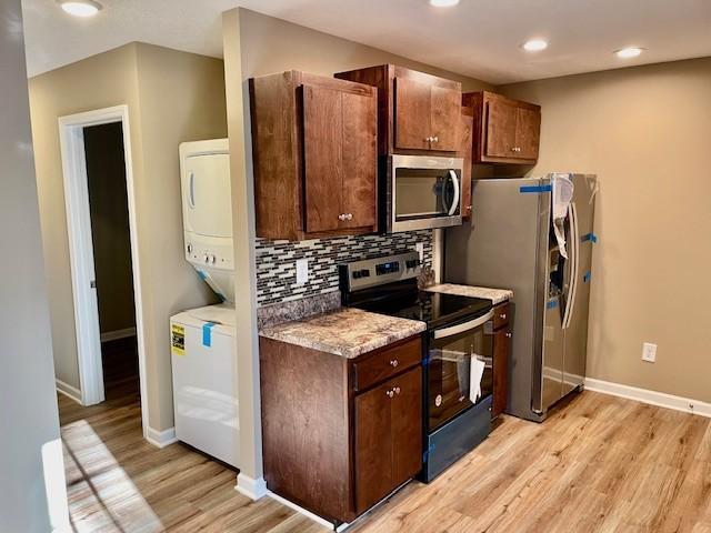 kitchen featuring tasteful backsplash, light wood-type flooring, stainless steel appliances, stacked washer and clothes dryer, and baseboards