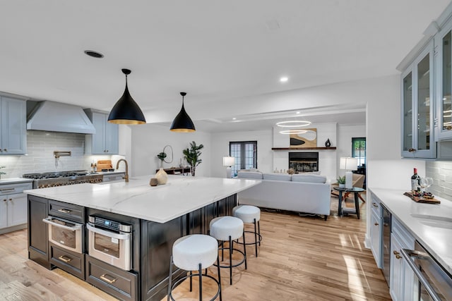 kitchen with custom exhaust hood, a kitchen island with sink, hanging light fixtures, light wood-type flooring, and stainless steel appliances