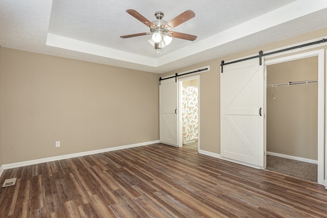 unfurnished bedroom with ceiling fan, a raised ceiling, dark hardwood / wood-style flooring, a barn door, and a closet