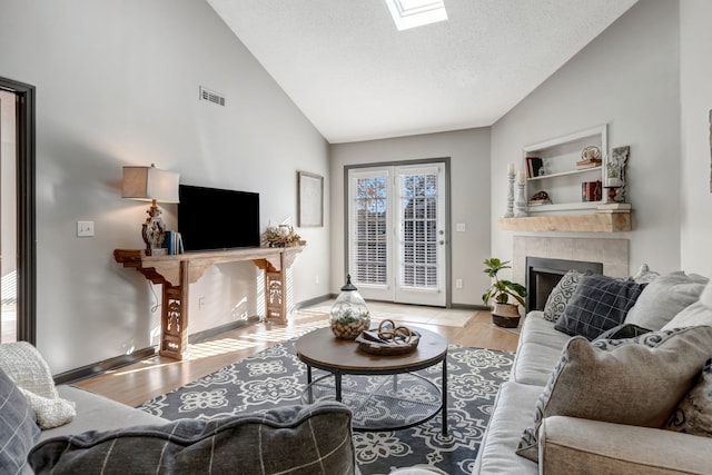 living room featuring a skylight, a textured ceiling, high vaulted ceiling, light hardwood / wood-style floors, and a tiled fireplace