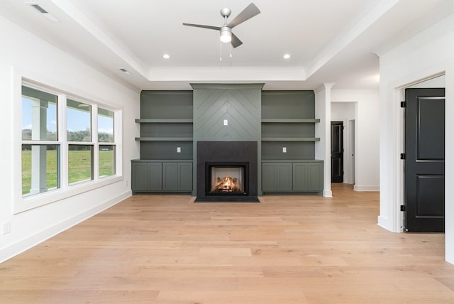 unfurnished living room featuring light wood-type flooring, a large fireplace, a raised ceiling, ceiling fan, and built in features